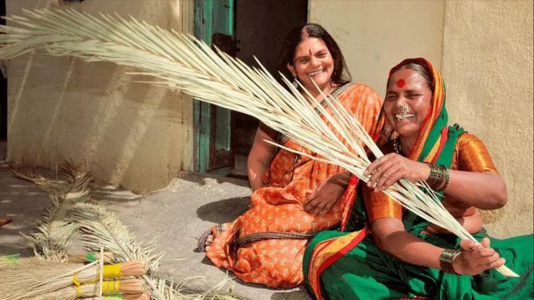 Chetna Gala Sinha, Founder Of India'S First Rural Women'S Cooperative Bank, Sitting On The Floor Outside A House In A Village, Wearing An Orange Saree. She Is Seen Laughing Along With A Traditional Rural Woman Who Is Sitting Next To Her In A Green Saree With Orange Blouse And Making Broomsticks. 