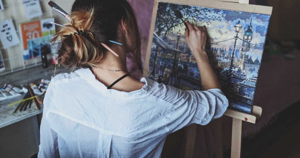 A Woman Painting A Scenery Of A House And Lake
